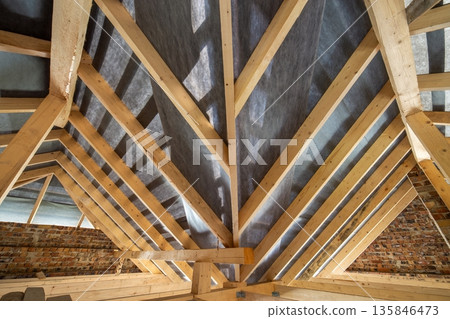 Attic of a building under construction with wooden beams of a roof structure and brick walls. Attic of a building under construction with wooden beams of a roof structure and brick walls. 135846473