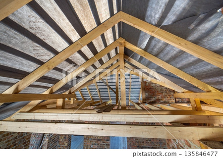 Attic of a building under construction with wooden beams of a roof structure and brick walls. Attic of a building under construction with wooden beams of a roof structure and brick walls. 135846477