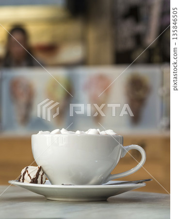 Mug of coffee with marshmallows on porcelain plate on blurred colorful interior bokeh background. Mug of coffee with marshmallows on porcelain plate on blurred colorful interior bokeh background. 135846505