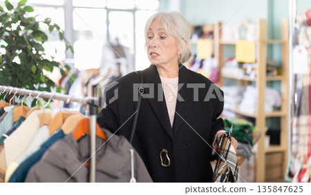 Elegance mature lady choosing warm coat or jacket for winter season in clothing boutique Elegance mature lady choosing warm coat or jacket for winter season in clothing boutique 135847625