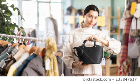 Fashionable young girl customer standing near shelves with clothes in store and choosing handbag 135847637