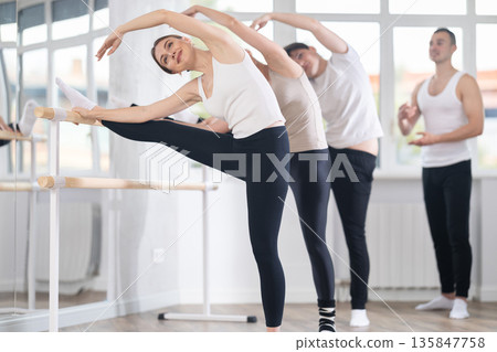 Beautiful athletic girl with group of dancers practicing ballet pose grand batman near ballet barre in dancing hall under guidance of experienced choreographer Beautiful athletic girl with group of dancers practicing ballet pose grand batman near ballet barre in dancing hall under guidance of experienced choreographer 135847758