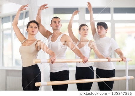 Warm-up sessions, young women and men doing ballet exercise standing in 3rd position in hall with ballet bar Warm-up sessions, young women and men doing ballet exercise standing in 3rd position in hall with ballet bar 135847856