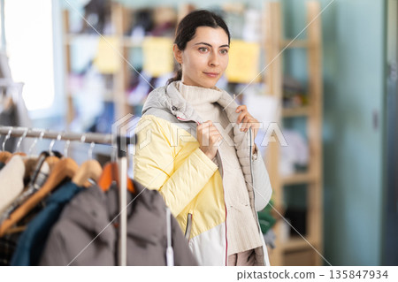 Shopping and winter sales concept. Focused young girl client choosing and trying on down jacket in clothing mall Shopping and winter sales concept. Focused young girl client choosing and trying on down jacket in clothing mall 135847934