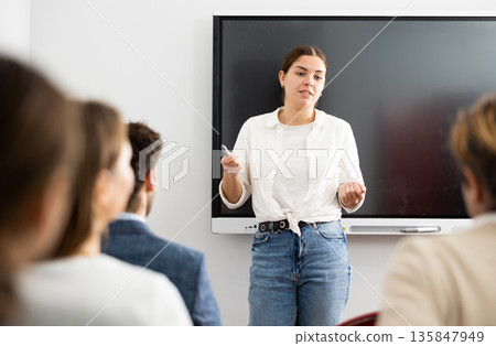 Portrait of young businesswoman standing near interactive board and participating in corporate seminar in auditorium Portrait of young businesswoman standing near interactive board and participating in corporate seminar in auditorium 135847949