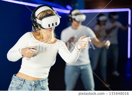 In darkened hall of gaming club, girl in VR helmet takes part in group game 135848016