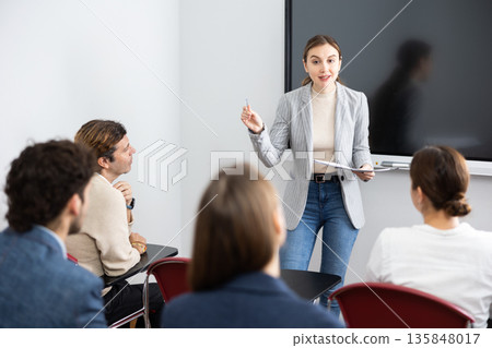 Positive skilled female coach explaining topic to group of people during foreign language class in auditory Positive skilled female coach explaining topic to group of people during foreign language class in auditory 135848017