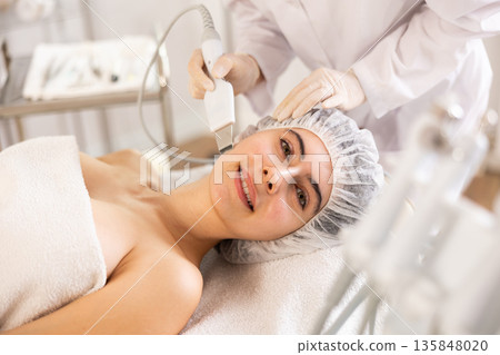 Young female client experiencing facial skin peeling using ultrasonic scrubber 135848020