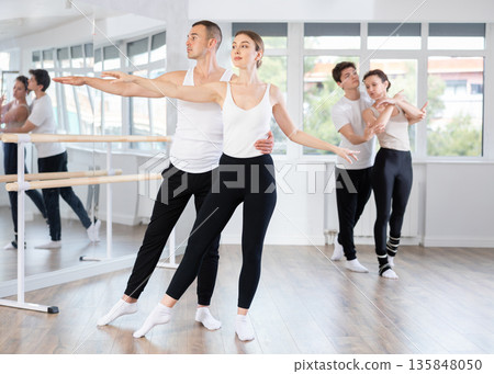 Smiling young man and female partner in black and white clothing with group active people training ballet dance movements in dance salon Smiling young man and female partner in black and white clothing with group active people training ballet dance movements in dance salon 135848050