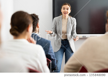 Young woman standing near interactive board and communicating with adult students during advanced training courses in classroom 135848090
