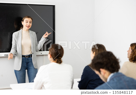 Young female teacher near interactive board, gesturing with hand in front of people sitting at desks in auditory Young female teacher near interactive board, gesturing with hand in front of people sitting at desks in auditory 135848117