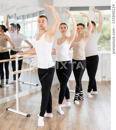 Experienced young male ballet dancer, dressed in white and black and group of girls and guys engaged in classical ballet performs exercise standing in 3rd position near ballet barre in dance hall 135848124