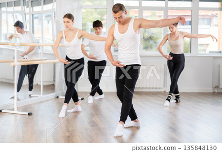 Experienced young male ballet dancer, dressed in white and black executing dance moves with focused precision among group of people rehearsing in ballet studio 135848130