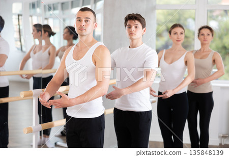 Sports young man in black and white sportswear, training first position of ballet stance at barre with group in choreography studio 135848139