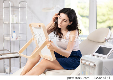 Young brunette woman admires her new face in mirror after plastic surgery Young brunette woman admires her new face in mirror after plastic surgery 135848141