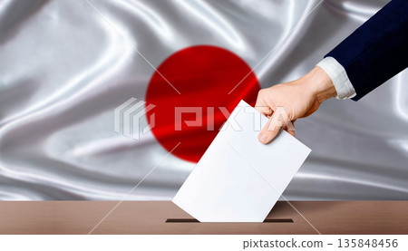 Hand placing a ballot in ballot box with the Japanese flag in the background. Japan presidential vote 2026 Hand placing a ballot in ballot box with the Japanese flag in the background. Japan presidential vote 2026 135848456