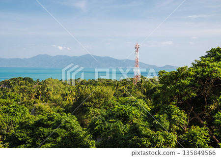 Telecommunication tower rising above tropical forest with turquoise sea, distant island mountains and wide blue sky, natural landscape 135849566