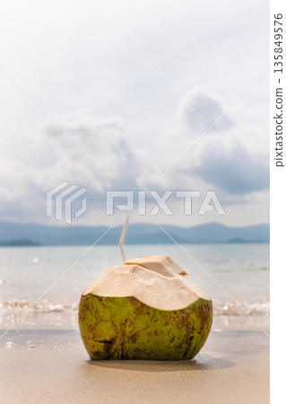 Fresh green coconut with straw on wet sandy beach, calm sea and cloudy sky in background, tropical summer vacation scene  135849576