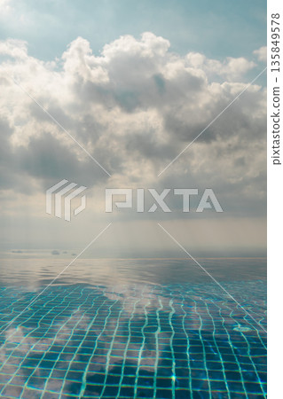 Calm infinity pool surface reflecting soft clouds and pale sunlight above distant sea creating minimal tropical background with open sky and tranquil resort atmosphere Calm infinity pool surface reflecting soft clouds and pale sunlight above distant sea creating minimal tropical background with open sky and tranquil resort atmosphere 135849578
