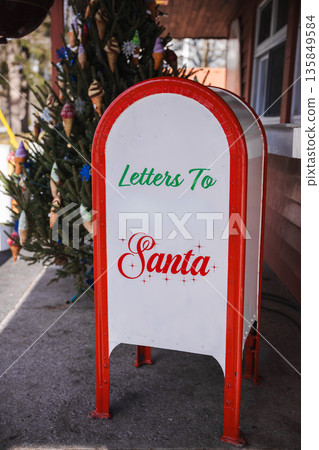 Vintage red and white mailbox for letters to Santa standing outdoors beside a decorated Christmas tree. 135849584