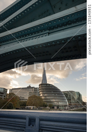 From beneath the imposing, riveted metalwork of Tower Bridge, the London skyline unfolds, featuring the distinct architecture of City Hall and the soaring spire of The Shard skyscraper in the From beneath the imposing, riveted metalwork of Tower Bridge, the London skyline unfolds, featuring the distinct architecture of City Hall and the soaring spire of The Shard skyscraper in the 135849931