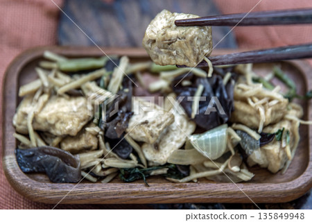 The hand is using chopsticks to pick up Stir-fried tofu with ginger in wooden tray. 135849948