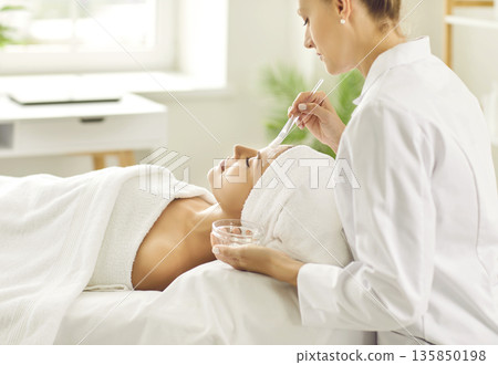 Professional beautician applying cleansing mask on face of young woman at spa salon Professional beautician applying cleansing mask on face of young woman at spa salon 135850198