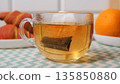 Close-up of a teabag dipping into hot water. Croissants and oranges in the background, calm morning tea and breakfast concept  135850880