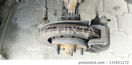 Close-up of a dirty, vented disc brake assembly with caliper, showing visible wear marks on the rotor and rusty wheel studs against a light gray paved background 135851272
