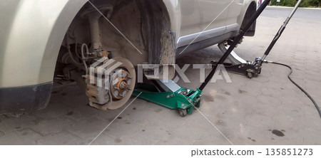 The beige car is lifted with a green jack, revealing a dusty rear disc brake, suggesting the wheel has been removed for repair or outdoor maintenance. The beige car is lifted with a green jack, revealing a dusty rear disc brake, suggesting the wheel has been removed for repair or outdoor maintenance. 135851273
