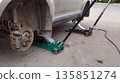Beige car is lifted by a green floor jack, exposing the dusty rear disc brake assembly, suggesting a wheel removal for repair or maintenance work outdoors 135851274