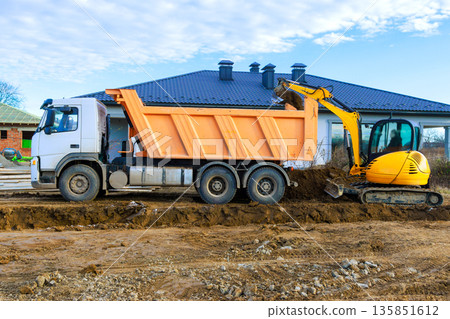 Dump truck is parked next to house while an excavator digs in dirt at construction site. 135851612