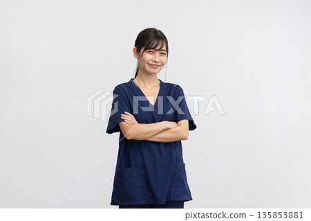 一位身穿藏藍色醫用工作服的年輕女子,面帶微笑,雙手交叉,站在白色背景前。 一位身穿藏藍色醫用工作服的年輕女子,面帶微笑,雙手交叉,站在白色背景前。 135853881