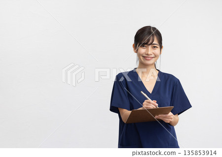 A young woman wearing navy blue scrubs and holding a binder and pen in front of a white background, smiling A young woman wearing navy blue scrubs and holding a binder and pen in front of a white background, smiling 135853914