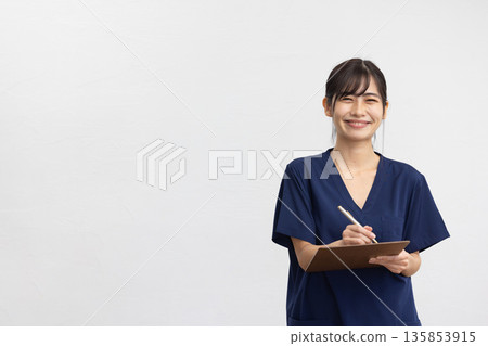 A young woman wearing navy blue scrubs and holding a binder and pen in front of a white background, smiling A young woman wearing navy blue scrubs and holding a binder and pen in front of a white background, smiling 135853915