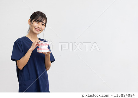 A young woman wearing navy blue medical scrubs smiling while holding a dental model in front of a white background A young woman wearing navy blue medical scrubs smiling while holding a dental model in front of a white background 135854084