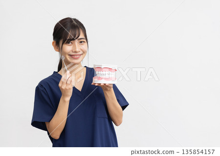 一位身穿藏藍色手術服的年輕女子麵帶微笑,手持牙科模型和牙科器械,站在白色背景前。 一位身穿藏藍色手術服的年輕女子麵帶微笑,手持牙科模型和牙科器械,站在白色背景前。 135854157