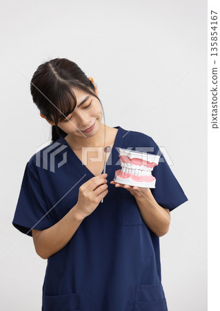 A young woman wearing navy blue scrubs smiling while holding a dental model and dental instruments in front of a white background A young woman wearing navy blue scrubs smiling while holding a dental model and dental instruments in front of a white background 135854167