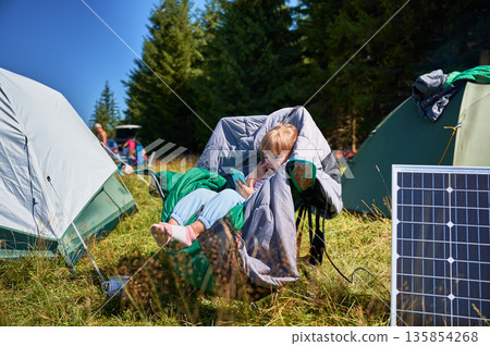 Young child plays with smartphone while charging with photovoltaic solar panel near tourist tent in summer. Integration of renewable energy in outdoor camping activities. Young child plays with smartphone while charging with photovoltaic solar panel near tourist tent in summer. Integration of renewable energy in outdoor camping activities. 135854268
