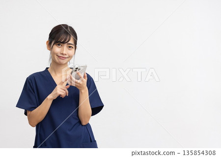 A young woman wearing a navy blue scrub and smiling at the camera with a smartphone in her hand in front of a white background A young woman wearing a navy blue scrub and smiling at the camera with a smartphone in her hand in front of a white background 135854308