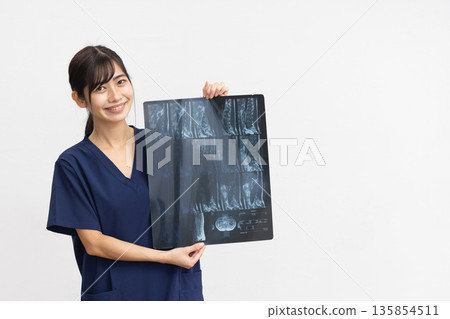 一位身穿藏藍色手術服的年輕女子麵帶微笑，手持X光片，站在白色背景前。 135854511
