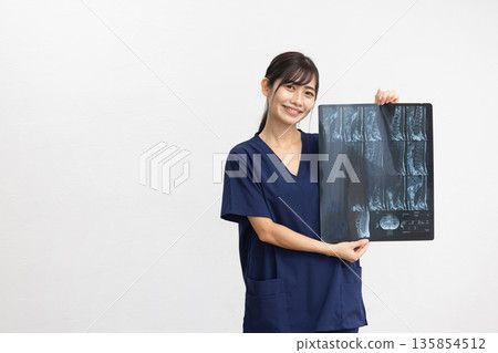A young woman wearing a navy blue scrub smiling while holding an X-ray in front of a white background A young woman wearing a navy blue scrub smiling while holding an X-ray in front of a white background 135854512