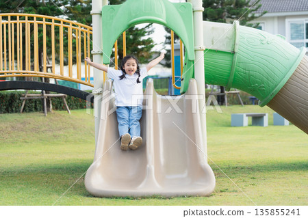 Happy child sliding down playground slide with arms raised, enjoying outdoor playtime on sunny day in community park surrounded by green grass Happy child sliding down playground slide with arms raised, enjoying outdoor playtime on sunny day in community park surrounded by green grass 135855241
