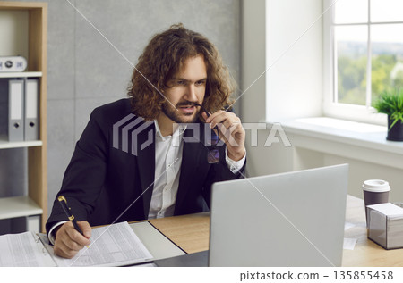 Young business man working on a laptop at office and looking on a computer screen. Young business man working on a laptop at office and looking on a computer screen. 135855458