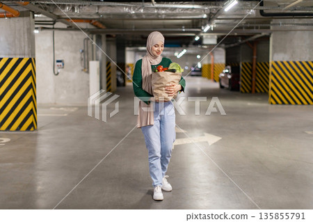 A woman wearing a hijab carries a bag of fresh groceries while walking through a parking garage A woman wearing a hijab carries a bag of fresh groceries while walking through a parking garage 135855791