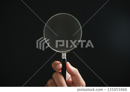 Hand holding a magnifying glass against a black background. The glass is slightly dusty. Hand holding a magnifying glass against a black background. The glass is slightly dusty. 135855988