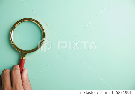Hand holding magnifying glass with gold rim on turquoise background. 135855991