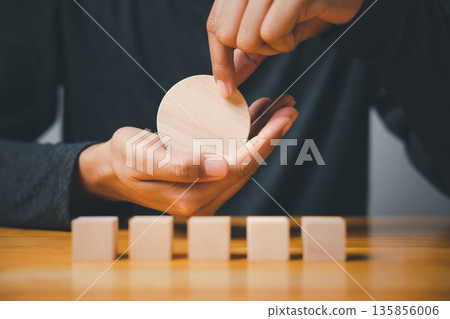 Hand holding wooden circle over a row of wooden blocks on a table. 135856006