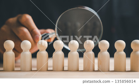 A hand holding a magnifying glass over a row of wooden figurines. The magnifying glass focuses on one figurine, emphasizing the importance of individual selection. A hand holding a magnifying glass over a row of wooden figurines. The magnifying glass focuses on one figurine, emphasizing the importance of individual selection. 135856014