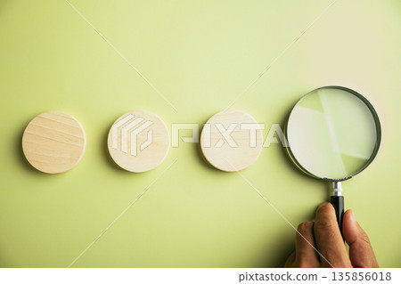 Three wooden circles arranged in a row, with a magnifying glass positioned to the right.  A hand holds the magnifying glass.  The background is a light green. 135856018